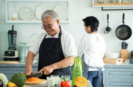 An aged man cutting vegetables while a woman is cooking in the background